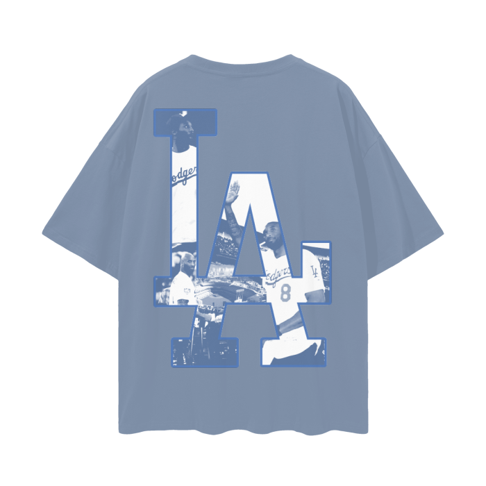 A blue oversized t-shirt featuring a large "LA" logo on the back, filled with images of Kobe Bryant wearing a Dodgers jersey. The collage-style design integrates various iconic moments of Kobe in the Dodgers attire, creating a tribute to his connection with Los Angeles sports culture.