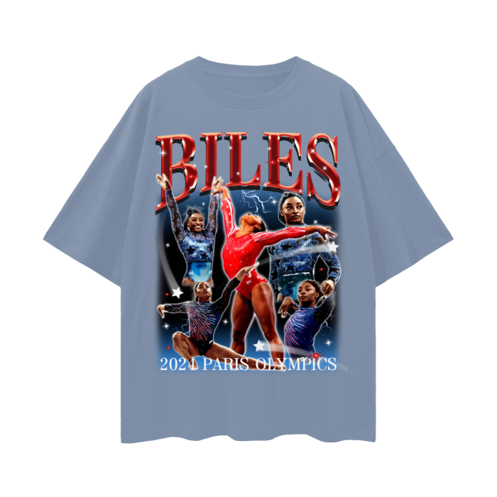 A light grey blue oversized t-shirt featuring a vibrant collage of Simone Biles in various gymnastic poses. The design prominently displays the name "BILES" in bold red letters at the top and includes the text "2024 Paris Olympics" at the bottom.
