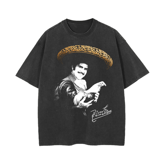 A black vintage-style T-shirt featuring a grayscale image of Vicente Fernandez holding a rooster. The design includes a large sombrero with ornate gold detailing above his head and his signature below. The T-shirt has a worn, vintage appearance
