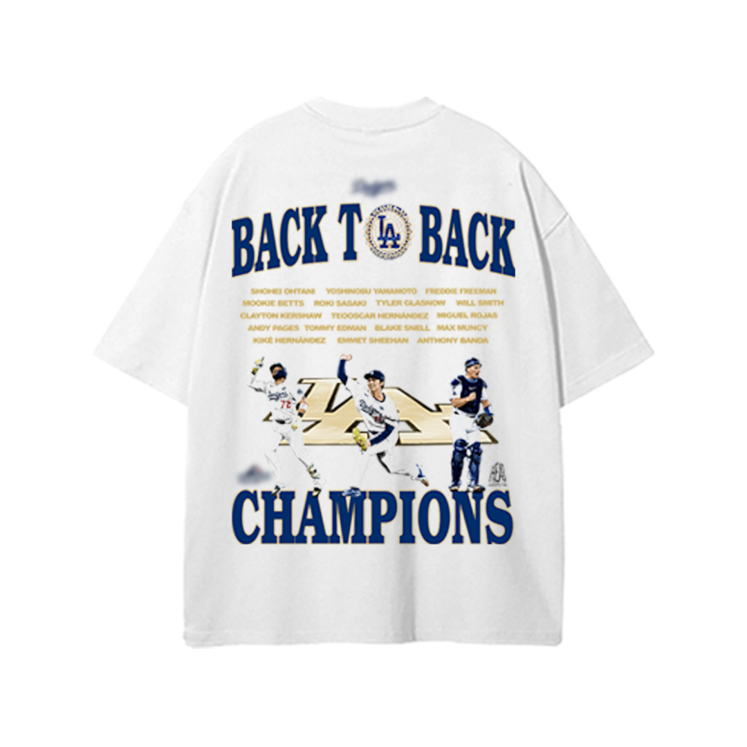 White oversized T-shirt featuring a bold graphic design celebrating the Los Angeles Dodgers’ back-to-back championships. The back of the shirt showcases the phrase “BACK TO BACK CHAMPIONS” in large navy and gold block letters, with the LA Dodgers logo between “BACK TO BACK.” Below, a lineup of players in action poses is positioned above the word “CHAMPIONS.” Above the graphic, the full roster names are printed in small gold text.