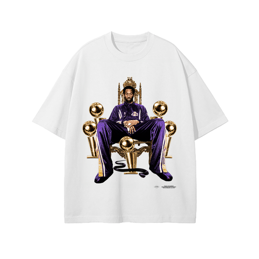 A white T-shirt featuring an image of Kobe Bryant sitting on a golden throne with basketball trophies surrounding him. He is dressed in a purple tracksuit, wearing a crown, and the design highlights his legacy as an NBA champion