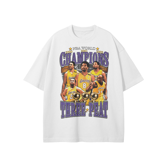 A white oversized T-shirt featuring the Los Angeles Lakers’ three-peat NBA championships. The front design includes portraits of key players like Kobe Bryant and Shaquille O'Neal with the text "NBA World Champions Three-Peat" in purple and gold.