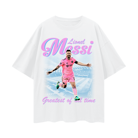 A white oversized Lionel Messi soccer tee showing Messi in his pink Inter Miami jersey with the text "Lionel Messi - Greatest of All Time" written in pink and purple font.