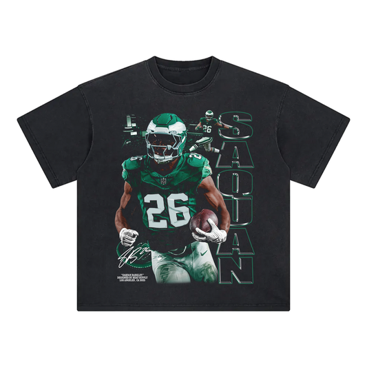 Black washed oversized T-shirt featuring a bold graphic of Saquon Barkley in a Philadelphia Eagles uniform, running with the football. The number 26 is displayed on his jersey, with his name 'Saquon' written vertically in bold green text alongside action shots of him in the background. The design includes a signature-style text and high-energy elements.